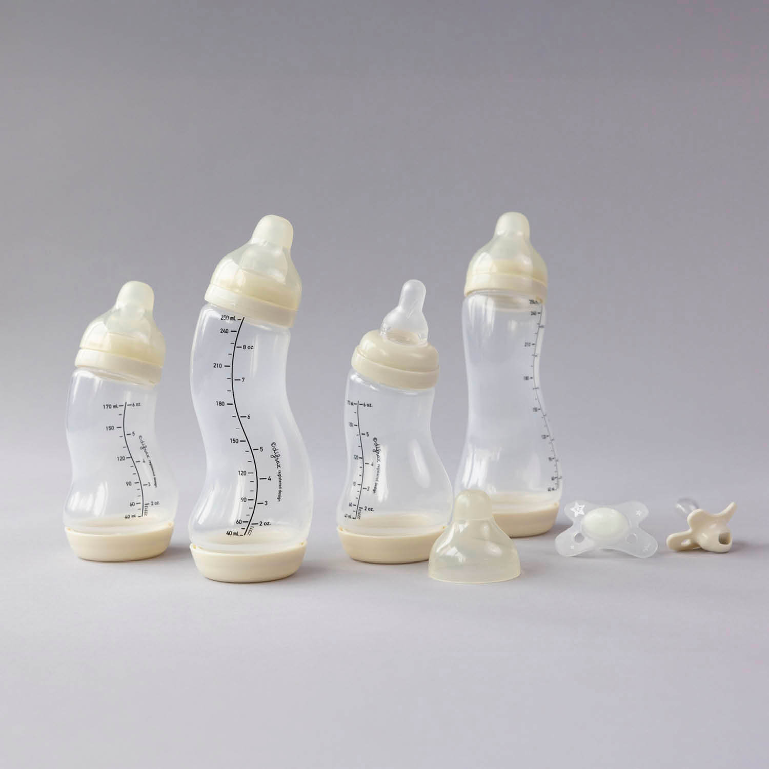 Difrax Newborn Starter Set compleet uitgepakt: 4 S-flesjes in ivory en 2 transparante fopspenen op grijze achtergrond