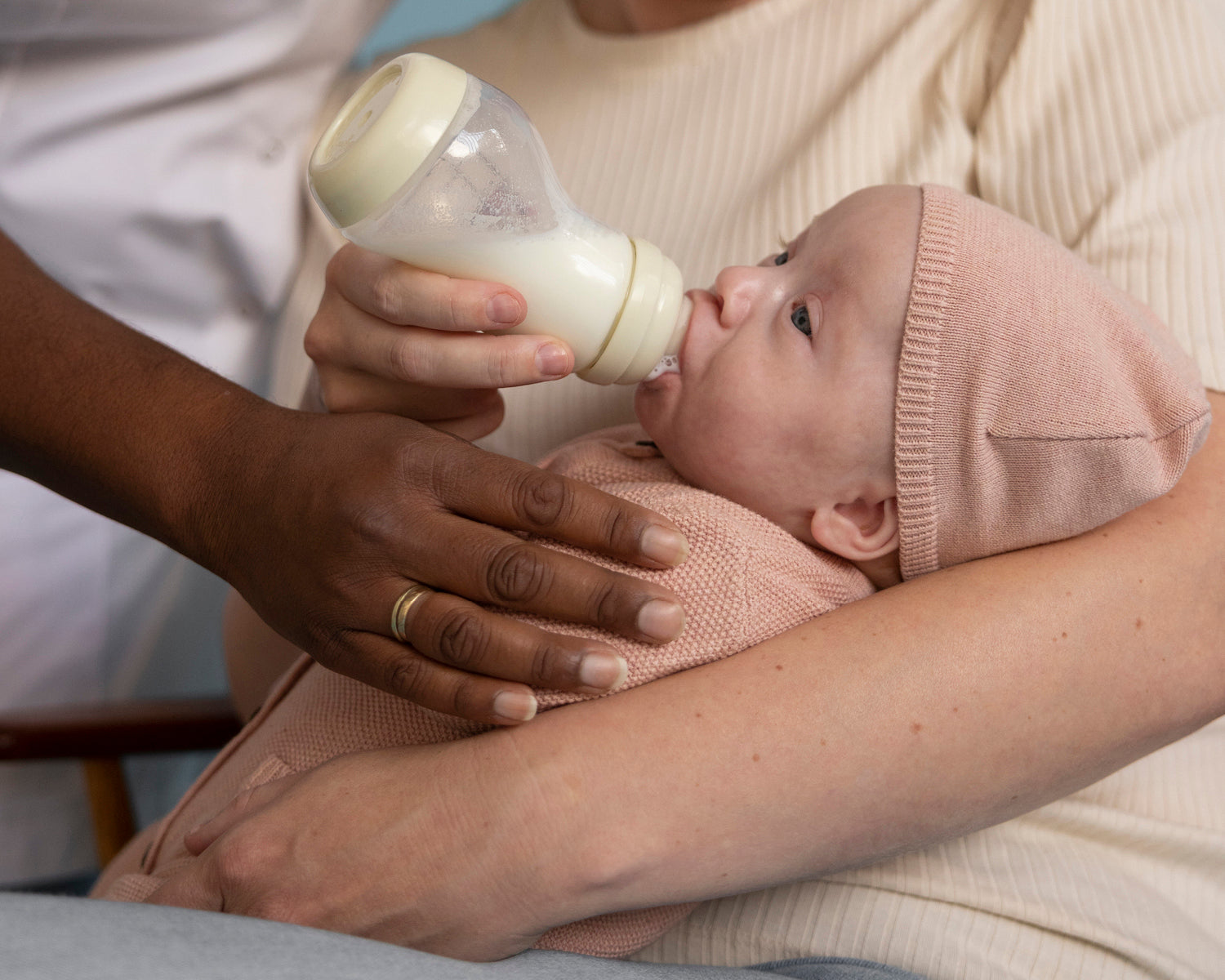 Kraamverzorgende voedt pasgeboren baby in roze mutsje met beige Difrax Wide S-fles in liefdevolle houding