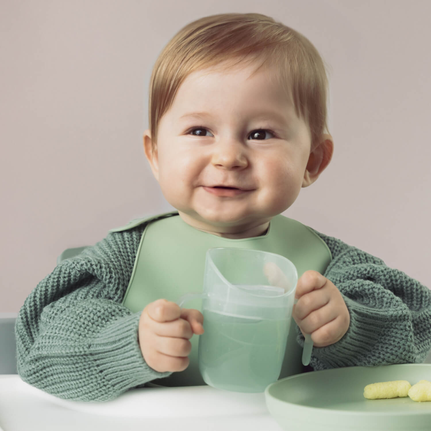 Dreumes zit aan de eettafel en houdt de Difrax LOVI Genius oefenbeker in pistachekleur vast, klaar om zelfstandig te drinken