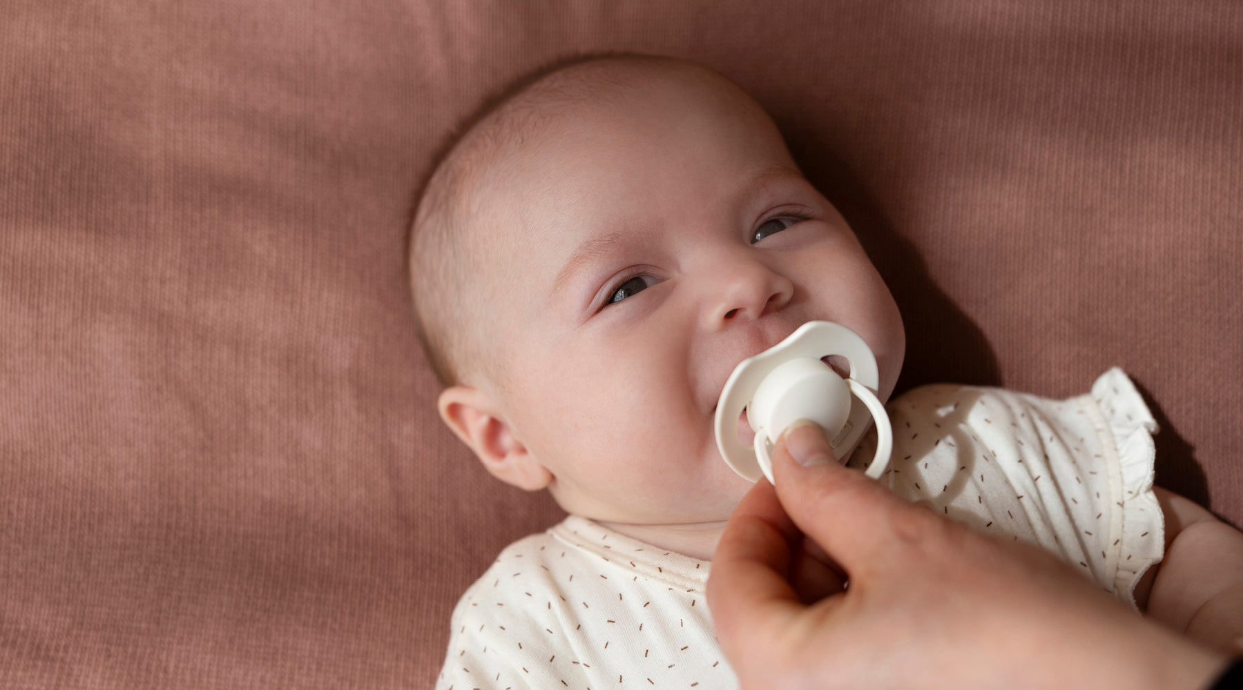 Baby in wit rompertje ligt op roze deken met witte fopspeen in de mond terwijl een hand de speen vasthoudt