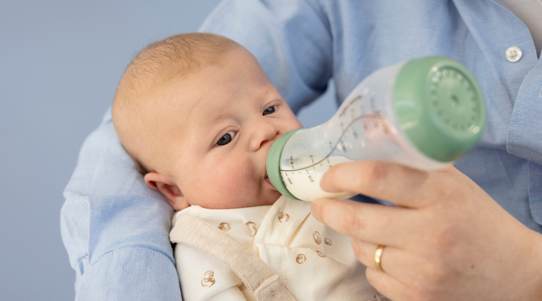 Pasgeboren baby drinkt uit mintgroene glazen Difrax S-fles met maatschaal in armen van ouder in blauw shirt