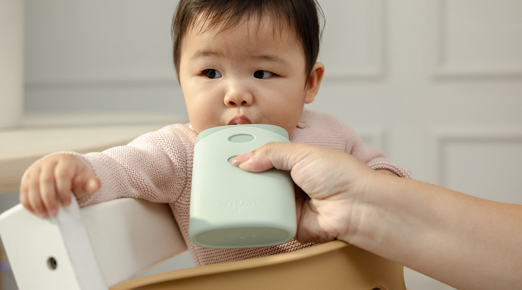 Baby in kinderstoel drinkt zelfstandig uit een knijpzakje met een knijpzakjeshouder, vastgehouden door een ouder