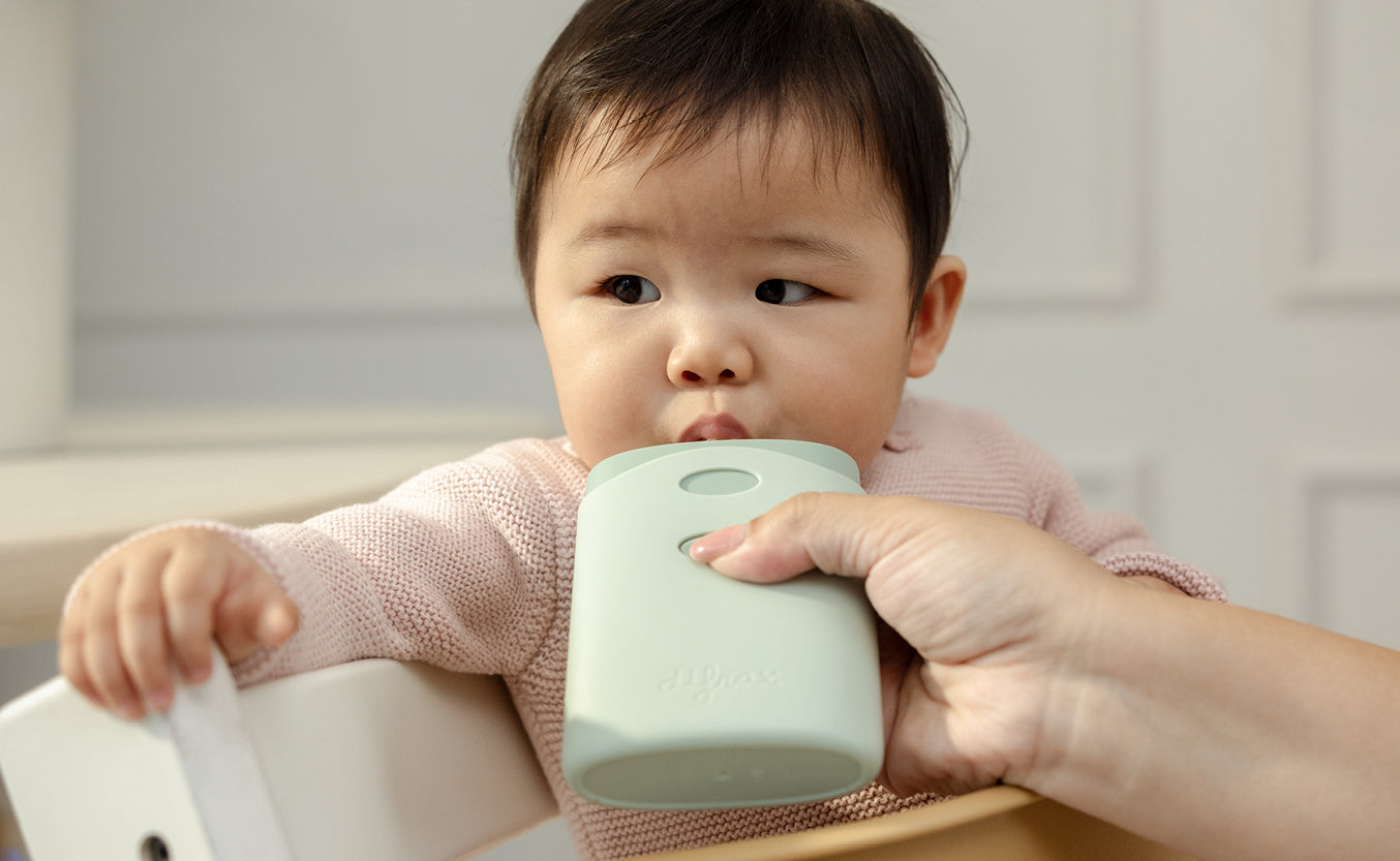 "Baby in roze gebreide trui drinkt uit een mintgroene siliconen pouchhouder die door een volwassene wordt vastgehouden