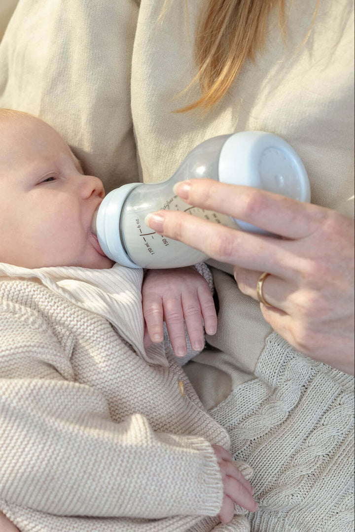 Moeder voedt pasgeboren baby met ijsblauwe Difrax S-fles in comfortabele voedingspositie, close-up van het voedingsmoment