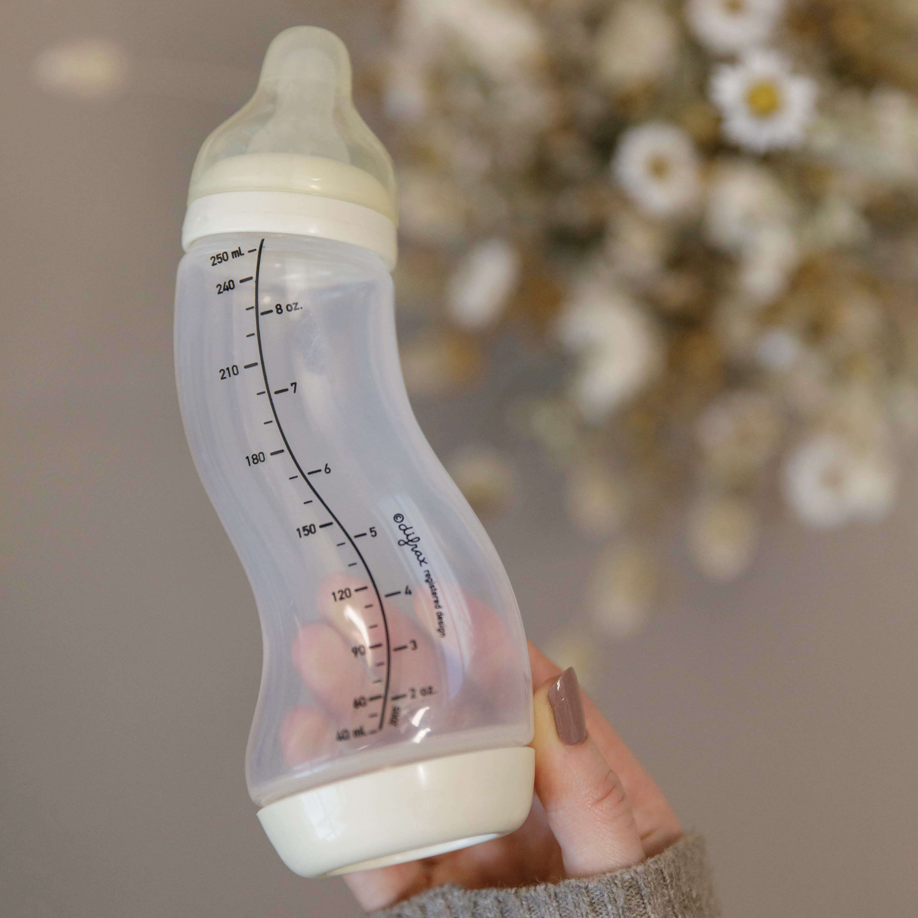 Hand houdt ivoorkleurige Difrax S-fles 250ml vast met droogbloemen op achtergrond, zachte neutrale tinten en natuurlijke setting