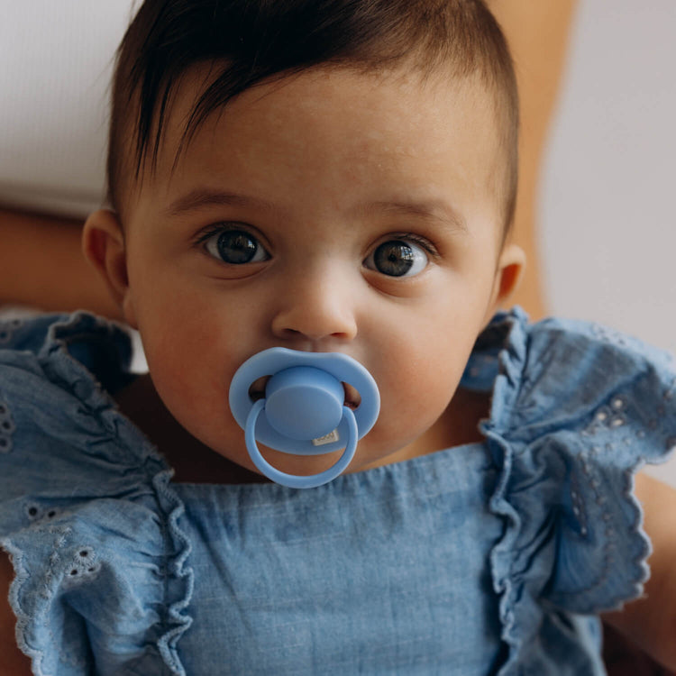 Donkerharige baby in een blauw jurkje met een lichtblauwe Difrax LOVI Dynamic fopspeen (Tutti Frutti, Ultramarine Blue)