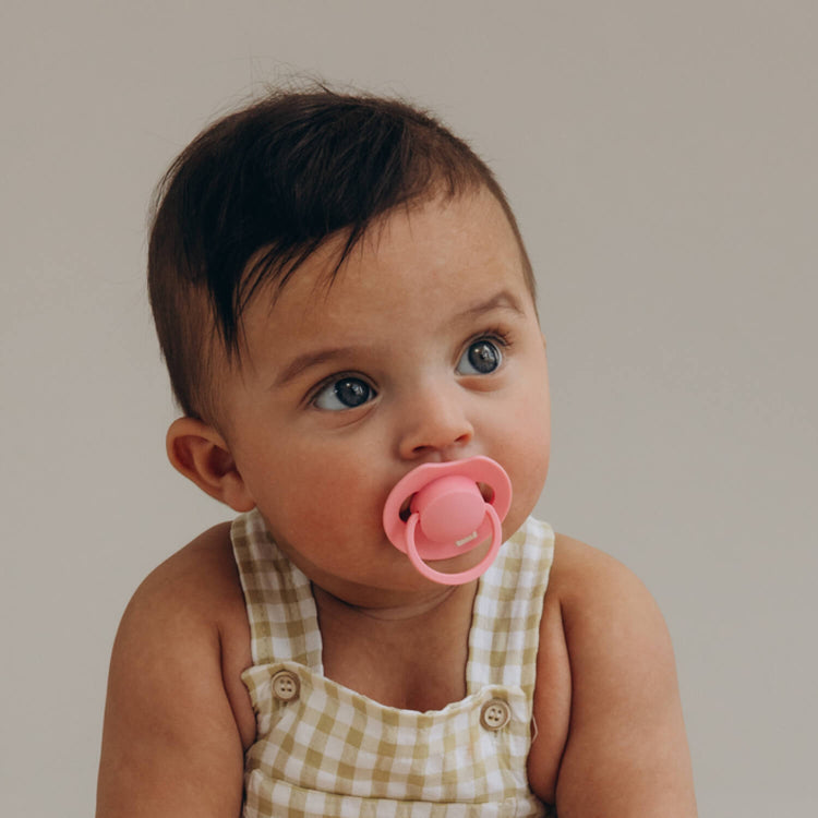 Donkerharige baby in een geruite tuinbroek met een roze Difrax LOVI Dynamic fopspeen (Tutti Frutti, Peony Pink)