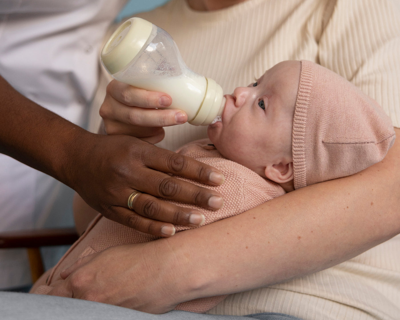Kraamverzorgende voedt pasgeboren baby in roze mutsje met beige Difrax Wide S-fles in liefdevolle houding
