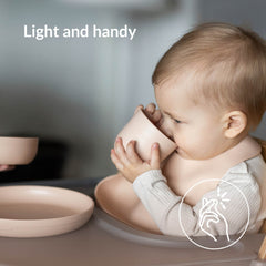 Baby drinkt uit de open beker met slabbetje en bord erbij – Difrax LOVI Baby Eet- & Drinkset Nude, lichtgewicht en handig in gebruik.